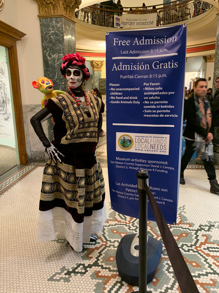 A woman in Dia de los Muertos face paint and traditional dresses poses in front of a welcome sign at the San Mateo County History Museum