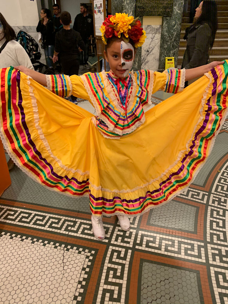 A young girl in Dia de los Muertos face paint and a traditional dress poses at the San Mateo County History Museum