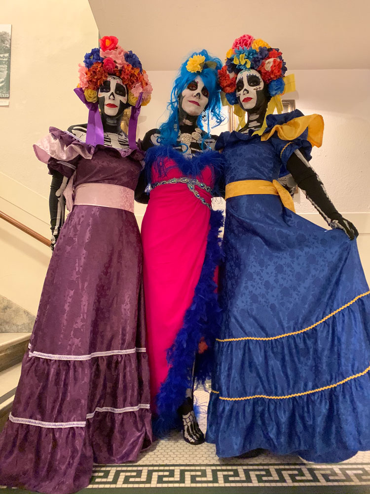 Three women in Dia de los Muertos face paint and traditional dresses poses at the San Mateo County History Museum