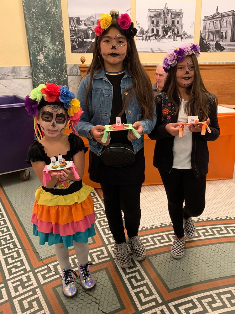 Three girls in Dia de los Muertos face paint and traditional dresses poses at the San Mateo County History Museum