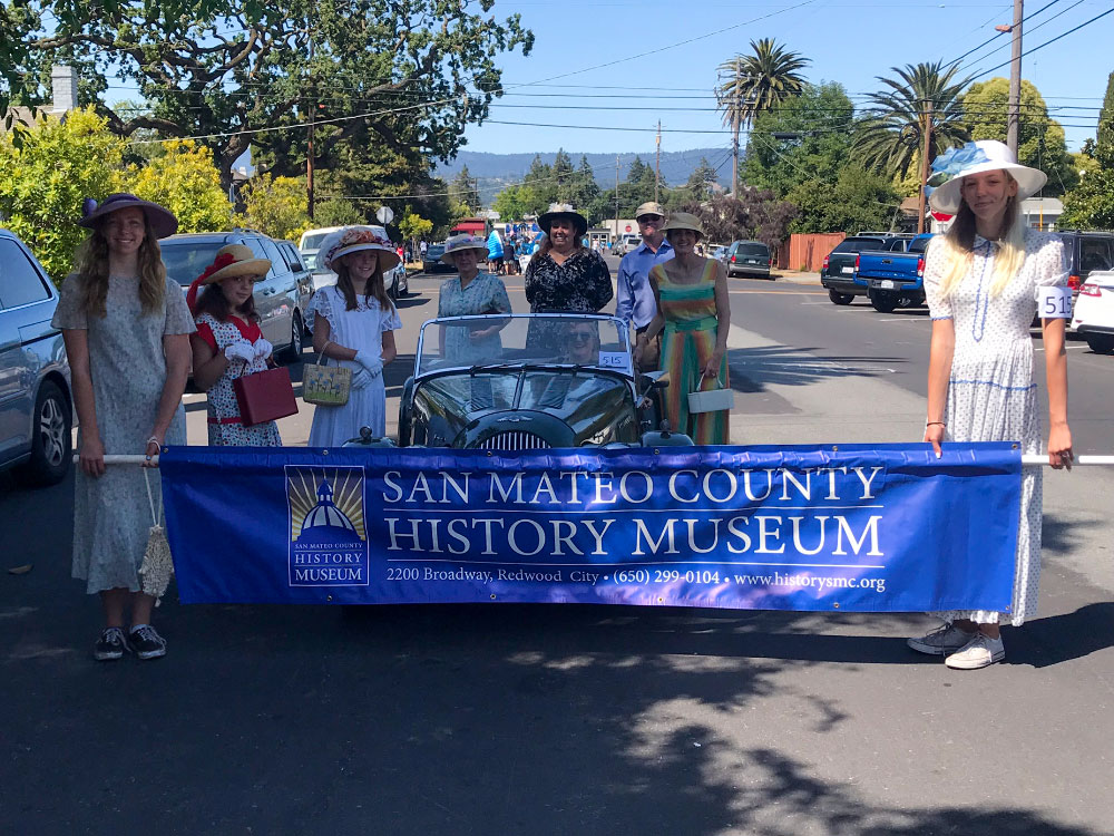 Parade gatherers in period clothing and a vintage care holding a sign for the San Mateo County History Museum