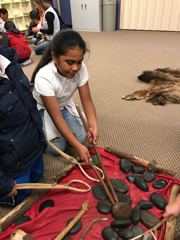 Young girl uses two sticks to pick up a stone at Providing Plenty school program at the San Mateo County History Museum