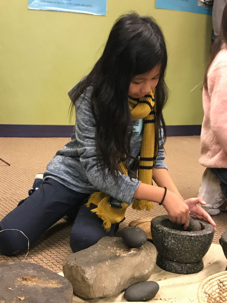 Young girl uses a mortar to ground acorns at Providing Plenty school program at the San Mateo County History Museum