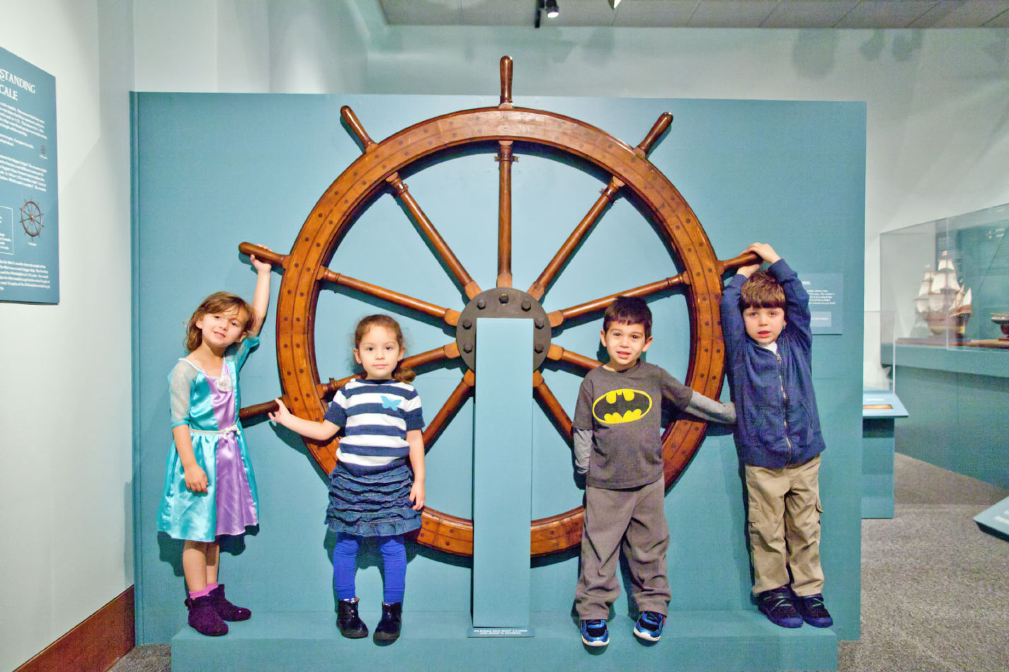 Children posing in front of a large ship steering wheel at Ships of the World exhibit at the San Mateo County History Museum