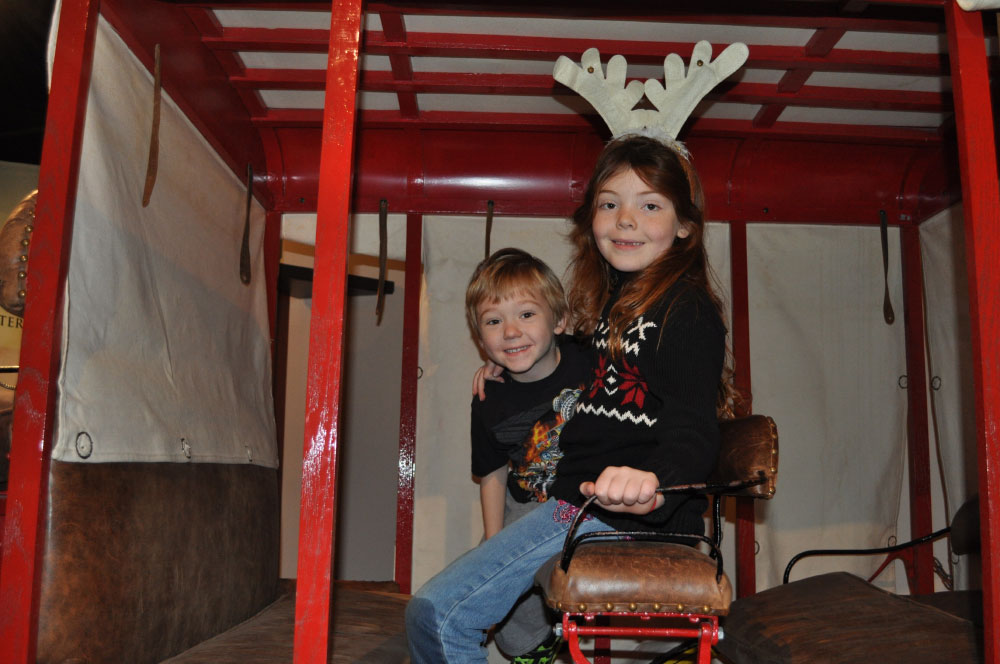 A young girl with festive antlers and young boy sitting in a carriage at Tree Treasures at the San Mateo County History Museum