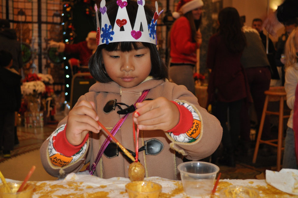 Young girl in holiday attire and paper crown making a tree ornament at the San Mateo History museum