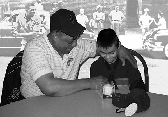 Willie Mays at a table with a child looking at autographed baseball