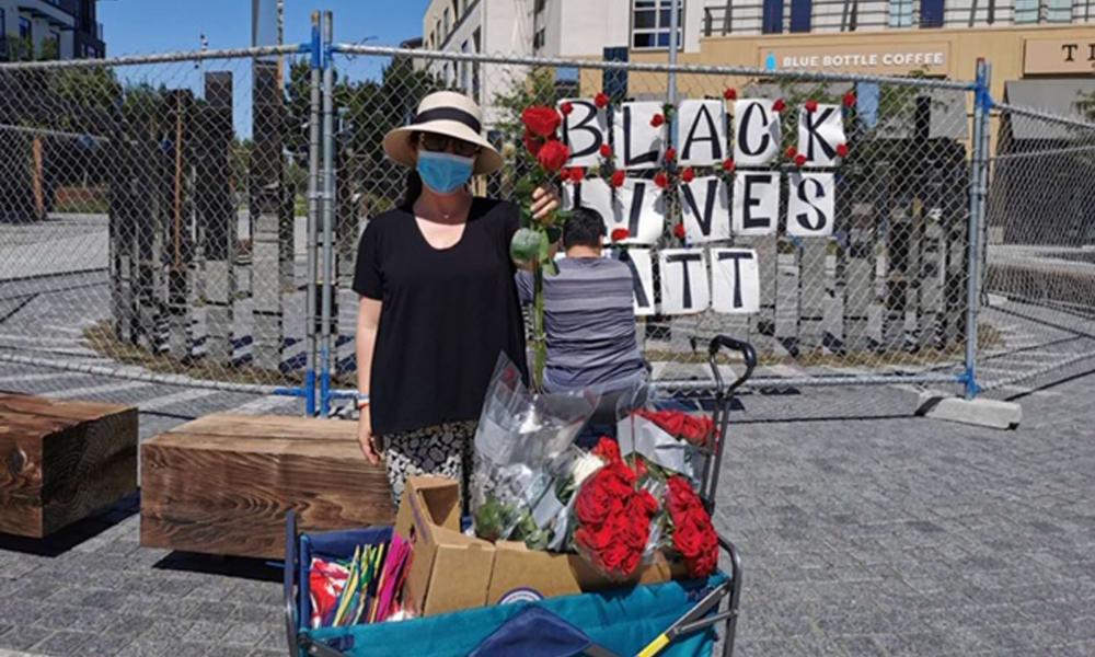 A woman wearing a mask for COVID 19 stands in front of a Black Lives Matter sign holding out roses