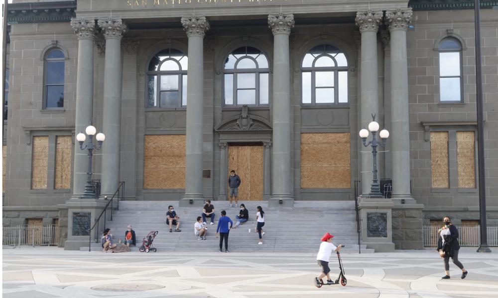 Entrance to the San Mateo County History Museum boarded up with people sitting on steps wearing masks due to COVID 19