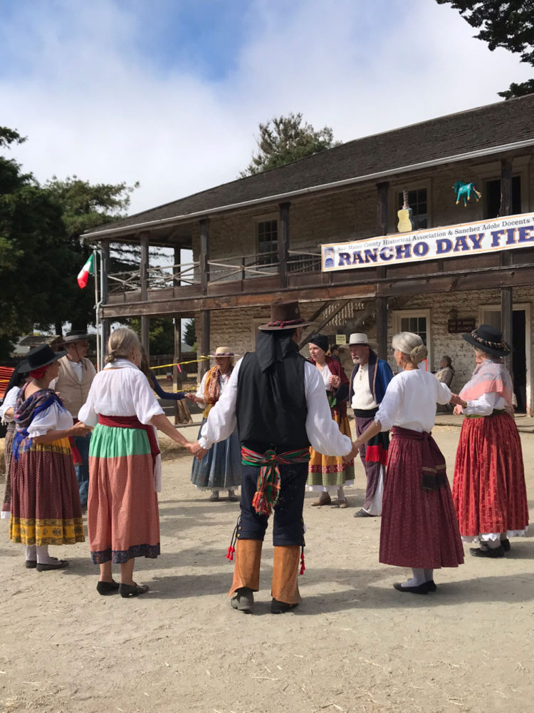 Guests hold hands in a circle in period costumes at the Sanchez Adobe Rancho Day Fiesta