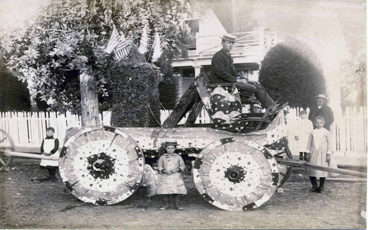 Family gathered around a Fourth of July float decorated with flags and flag bunting in front of a house circa 1880s