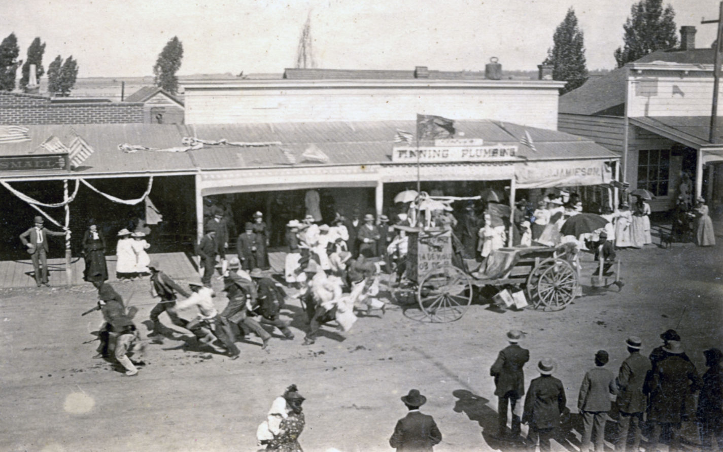Running men pull wagon down street during a parade circa 1880s