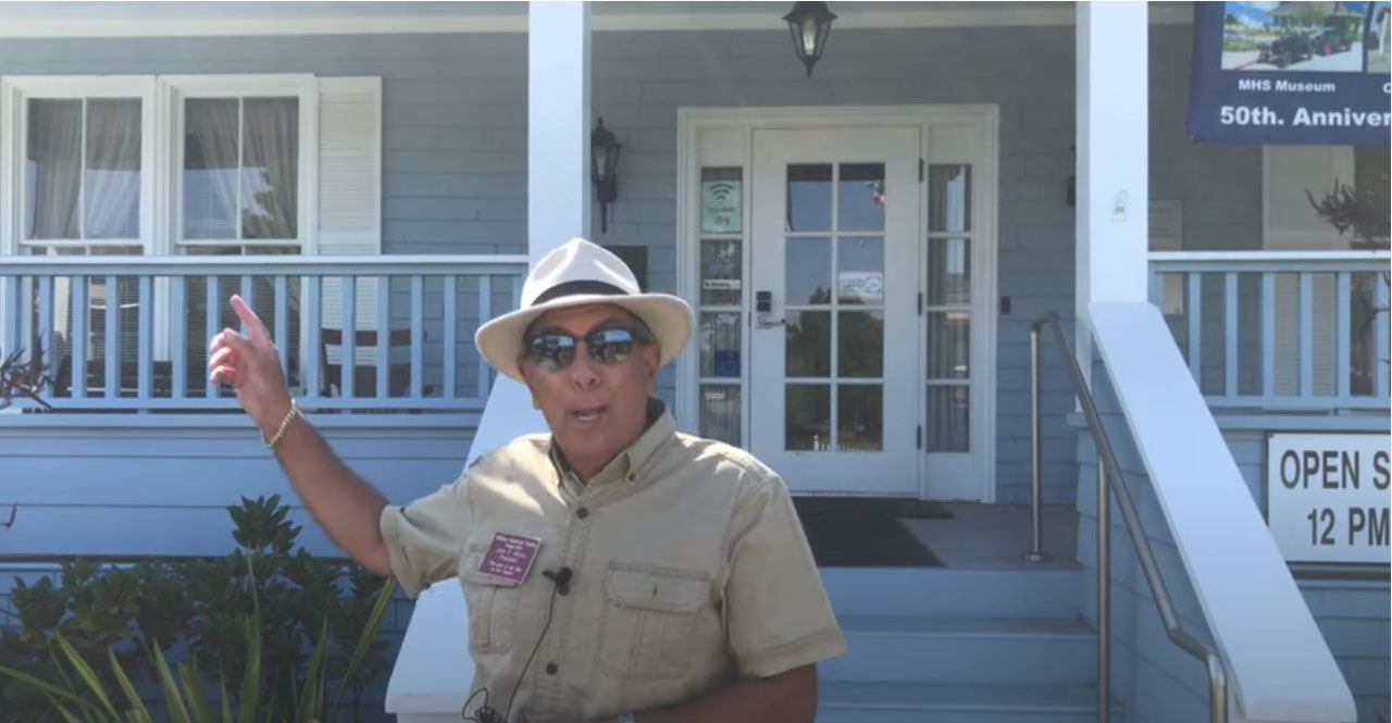 male docent in front of gray building with porch